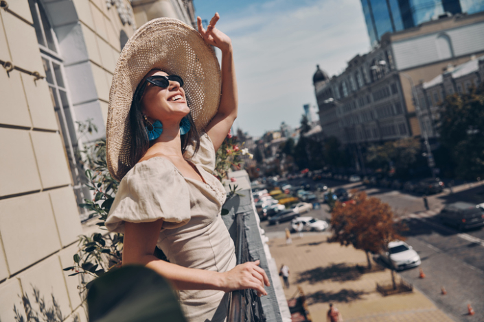 Relaxed young woman adjusting her elegant hat and smiling while standing on the balcony You see things as you are