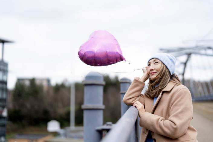 Woman with heart balloon stands by railing and looks up at cloudy sky in urban area
